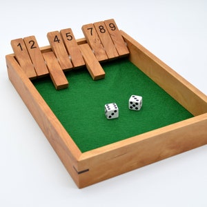 May include: A wooden Shut the Box game with a green felt playing surface. The game features wooden tiles numbered 1-9, and two white dice with black dots. The wooden box is rectangular.