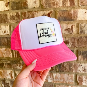 May include: Pink and white trucker hat with a white patch that says "Save Water Drink Champagne" with a pink champagne bottle graphic.