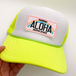 Könnte beinhalten: Eine weiße und neongelbe Trucker-Kappe mit einem Patch, auf dem "HAWAII ALOHA ALOHA STATE" steht, mit einem Regenbogen über dem Wort "ALOHA".