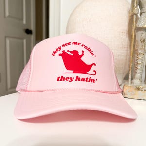 Puede incluir: Gorra de camionero rosa con un gráfico rojo de una persona montando en un trineo con el texto "they see me rollin' they hatin'".