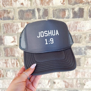 May include: A gray trucker hat with a black mesh back. The hat has white text that reads "JOSHUA 1:9".