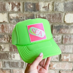 Könnte beinhalten: Eine neon-grüne Trucker-Kappe mit einem rosa quadratischen Patch, auf dem "SEA" in Form einer weißen Pille steht.