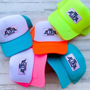 May include: A collection of trucker hats in various bright colors, including white, neon green, pink, orange, and teal. Each hat features a white patch with the word "ALOHA" and "HAWAII ALOHA STATE" in a rainbow design.