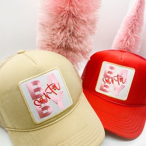Puede incluir: Dos gorras de camionero con un diseño de "Baby Santa" rosa y rojo. Las gorras son beige y rojas con una malla en la parte posterior.
