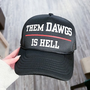 Puede incluir: Gorra de camionero negra con una franja roja y texto blanco que dice "Them Dawgs Is Hell".
