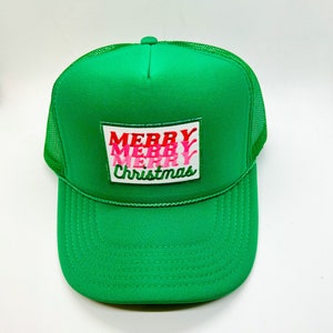 Puede incluir: Gorra de camionero verde con parte trasera de malla y visera curva. El panel frontal presenta un parche rectangular con las palabras "MERRY MERRY Christmas" en letras rojas y rosas.