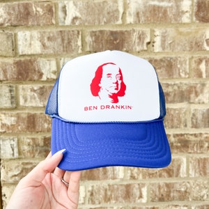 May include: A blue and white trucker hat with a red image of Benjamin Franklin and the text "BEN DRANKIN'" in red.