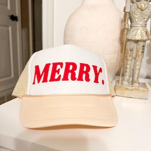 Puede incluir: Una gorra de camionero blanca y beige con un gráfico rojo "MERRY" en la parte delantera.