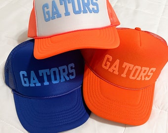 Puede incluir: Tres gorras de camionero con la palabra "GATORS" en letras azules en la parte delantera. Las gorras son naranja, blanca y azul.