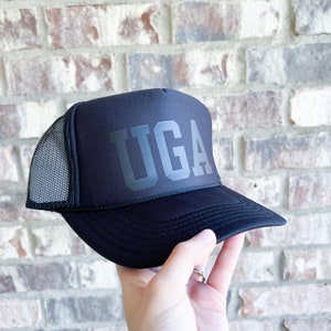 May include: Black trucker hat with a black mesh back and the word "UGA" in large, bold, black letters on the front.