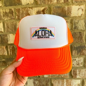 Puede incluir: Una gorra de camionero blanca y naranja con un arcoíris y el texto "HAWAII ALOHA ALOHA STATE" en un parche de placa de matrícula.