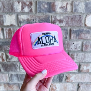 Puede incluir: Una gorra de camionero rosa con una parte trasera de malla. La gorra tiene un parche en la parte delantera que dice "HAWAII ALOHA ALOHA STATE" con un arcoíris sobre la palabra "ALOHA".