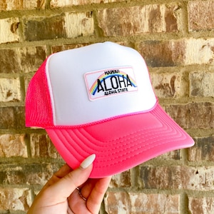 Puede incluir: Gorra de camionero rosa neón y blanca con visera curva. La gorra presenta un parche con la palabra "ALOHA" encima de "ALOHA STATE" y un diseño de arcoíris. La gorra tiene una parte trasera de malla y una parte delantera blanca.
