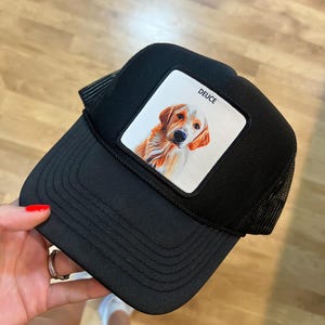Op de afbeelding: Zwarte truckerpet met een vierkante patch met een illustratie van een golden retriever. Het woord "DEUCE" staat boven de hond gedrukt. De pet heeft een mesh achterkant en een gebogen klep.