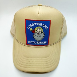 Op de afbeelding: Beige truckerpet met een mesh achterkant en een gebogen klep. De voorkant heeft een vierkante patch met de tekst "I DON'T BELIEVE IN YOU EITHER" en een cartoon illustratie van de kerstman.