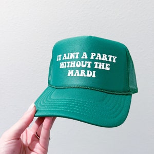 May include: Green trucker hat with the text "IT AINT A PARTY WITHOUT THE MARDI" in white. The hat has a mesh back and a curved brim. A hand is holding the hat.