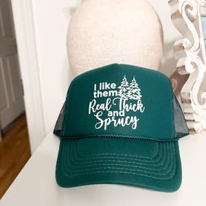 Puede incluir: Una gorra de camionero verde con una malla blanca en la parte posterior. La parte delantera de la gorra tiene un diseño blanco que dice "I like them Real Thick and Sprucy" con dos pinos estilizados.
