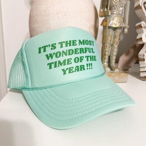 Puede incluir: Una gorra de camionero verde claro con una malla blanca en la parte posterior. La gorra tiene el texto "It's the most wonderful time of the year!!!" impreso en verde.