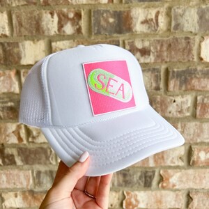 Könnte beinhalten: Weiße Trucker-Kappe mit einem rosa quadratischen Patch, auf dem eine grüne und weiße Pille mit dem Wort "SEA" gestickt ist.