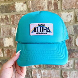 Puede incluir: Una gorra de camionero azul turquesa con una parte trasera de malla. La gorra tiene un parche en la parte delantera que dice "Hawaii Aloha Aloha State" con un arcoíris sobre el texto.