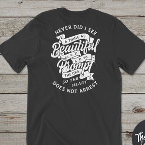Könnte beinhalten: Ein schwarzes T-Shirt mit einem weißen Grafikdesign, auf dem "Never did I see a thing as beautiful as a prompt to the heart so the heart does not arrest" steht. Die Grafik ist im Vintage-Stil mit einem Banner und einer Schriftart gehalten. Das T-Shirt hat auch ein kleines Logo unten rechts, auf dem "The Cath Lab Life" steht.