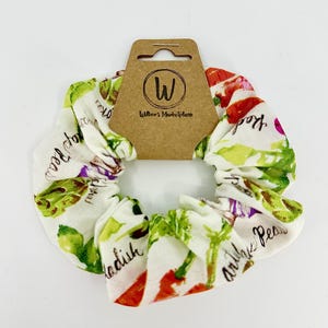 May include: White scrunchie with a colorful vegetable print featuring radishes, peas, and other produce. The scrunchie is paired with a brown tag that reads "Wilber's Marketplace" and features a "W" logo. A stylish hair accessory.