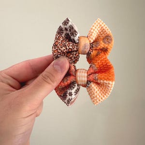 May include: A handmade hair bow with a mix of patterned fabrics. One side features a brown and tan leopard print, while the other side has an orange floral and gingham pattern. The bow is held together by a matching fabric center.