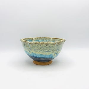 May include: A ceramic bowl with a scalloped rim featuring a reactive glaze in shades of cream, light blue, and deep navy. The exterior surface displays a speckled texture, while the base is finished in a contrasting unglazed tan clay.