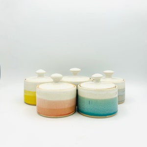 May include: Five ceramic lidded jars with a white glaze and colorful stripes. The jars are in a row, with the stripes going from yellow to pink to blue to gray.