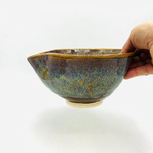 May include: A handmade ceramic bowl with a unique, organic shape. The bowl features a blend of brown, blue, and gold glaze, creating a textured, mottled appearance. The rim has a golden-brown edge, and the base is a lighter, unglazed color.