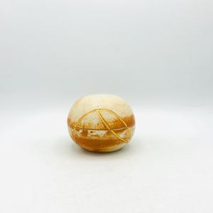 May include: A spherical ceramic lidded container featuring a cream-colored base with mottled orange-brown horizontal bands. The surface is accented with thin, raised gold-colored lines that form geometric patterns across the lid and body.
