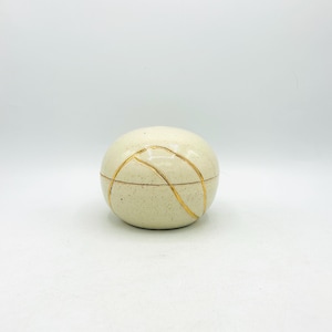 May include: A round, off-white ceramic lidded container with a speckled texture. The surface features thin, curved gold-colored lines that wrap around the exterior, crossing the seam where the lid meets the base of the small, spherical vessel.