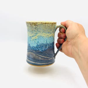 May include: A ceramic mug featuring a vertical gradient glaze that transitions from a pale yellow at the rim to a bright turquoise in the center and a deep navy blue at the base. The mug has a curved handle and a smooth, cylindrical body shape.