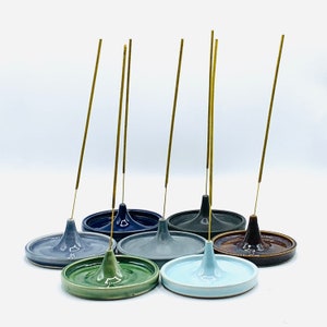 May include: Set of seven ceramic incense holders in various colors, including blue, green, brown, and gray. Each holder has a small cone-shaped indentation in the center and is designed to hold incense sticks. The holders are arranged in a semi-circle on a white surface.