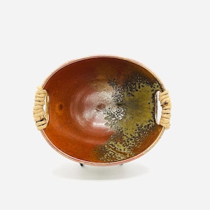 May include: An oval ceramic bowl featuring a deep reddish-brown interior that transitions into a textured, mottled olive-green glaze on one side. The bowl includes two integrated side handles wrapped in light tan natural fiber cordage.