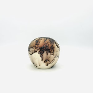 May include: A spherical ceramic vessel with a lid, featuring a smooth off-white base color and intricate, dark, web-like smoke firing patterns that wrap around the exterior surface, creating a unique contrast between the light and dark tones.
