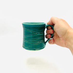 May include: A teal ceramic mug featuring a glossy finish and a series of carved, swirling horizontal lines that wrap around the exterior. The mug has a curved handle on the right side and a slightly flared rim, showcasing a deep, rich color.