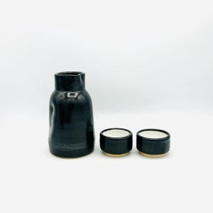 May include: A black ceramic sake set with a bottle and two small cups. The bottle has a unique, textured shape. The cups have a black exterior and a white interior, with a tan base. The set is on a white background.