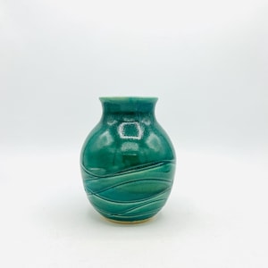 May include: A small, teal-colored ceramic vase with a glossy finish. The vase features a rounded body and a narrow neck. Horizontal wave-like patterns are carved into the surface, adding texture. The vase is suitable for displaying flowers or as a decorative item.