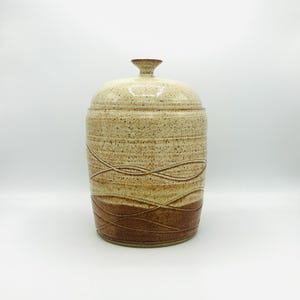 May include: A ceramic jar with a lid, featuring a speckled beige glaze and brown wavy lines. The jar has a rounded body and a slightly flared top.