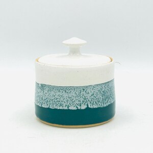 May include: A ceramic jar with a lid, featuring a white top, a teal-colored middle section with a textured, speckled appearance, and a dark teal base. The jar has a small, white knob on the lid.