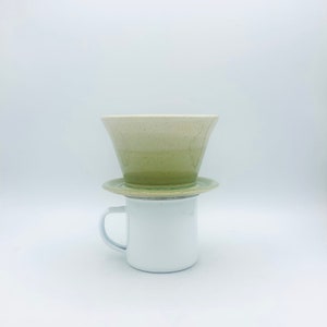 May include: A green and white ceramic pour-over coffee dripper with a white ceramic saucer sits on top of a white enamel mug.