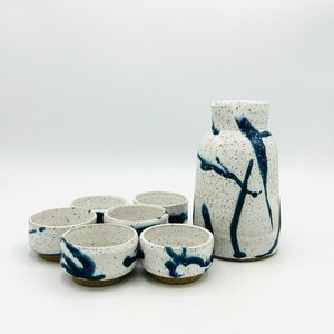 May include: A set of six white ceramic sake cups with blue abstract designs, and a matching white ceramic sake carafe with blue abstract designs.
