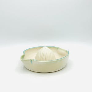 May include: A ceramic citrus juicer featuring a central ribbed reamer cone surrounded by a shallow basin. The piece has a creamy off-white glaze with subtle light blue accents along the rim and a small integrated pouring spout on one side.