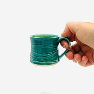 May include: A small ceramic mug with a glossy teal glaze and a textured surface featuring horizontal wavy lines. The mug has a curved handle on the right side and a smooth, rounded rim. The base of the mug shows a light beige, unglazed ceramic.