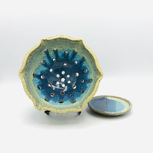 May include: A ceramic berry bowl with a scalloped rim, featuring a speckled yellow exterior and a deep blue interior with drainage holes. Beside it sits a matching small, round, flat ceramic saucer with a blue and yellow speckled glaze finish.