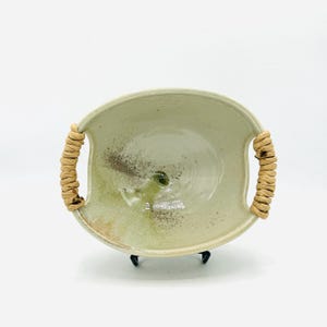 May include: A handmade ceramic bowl with a light green glaze and brown speckles. The bowl has two woven handles and sits on a black metal stand. The interior features a small, green, circular design.