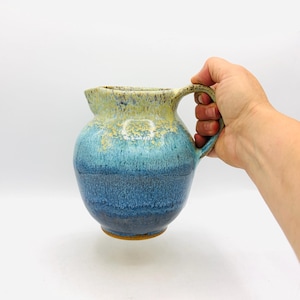May include: A ceramic pitcher featuring a rounded body and a curved handle. The glaze transitions from a pale yellow and cream color at the top rim to a vibrant turquoise in the middle, deepening into a dark navy blue at the base of the vessel.