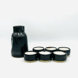 May include: A black ceramic sake set featuring a bottle and six small cups. The bottle has a textured surface and a narrow spout. The cups are round with a black exterior and a white interior.