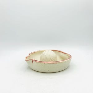 May include: A ceramic citrus juicer featuring a cream-colored bowl with a slightly irregular, organic rim and a central reamed dome. The edges of the bowl and the spout area are accented with a thin, textured reddish-brown glaze detail.